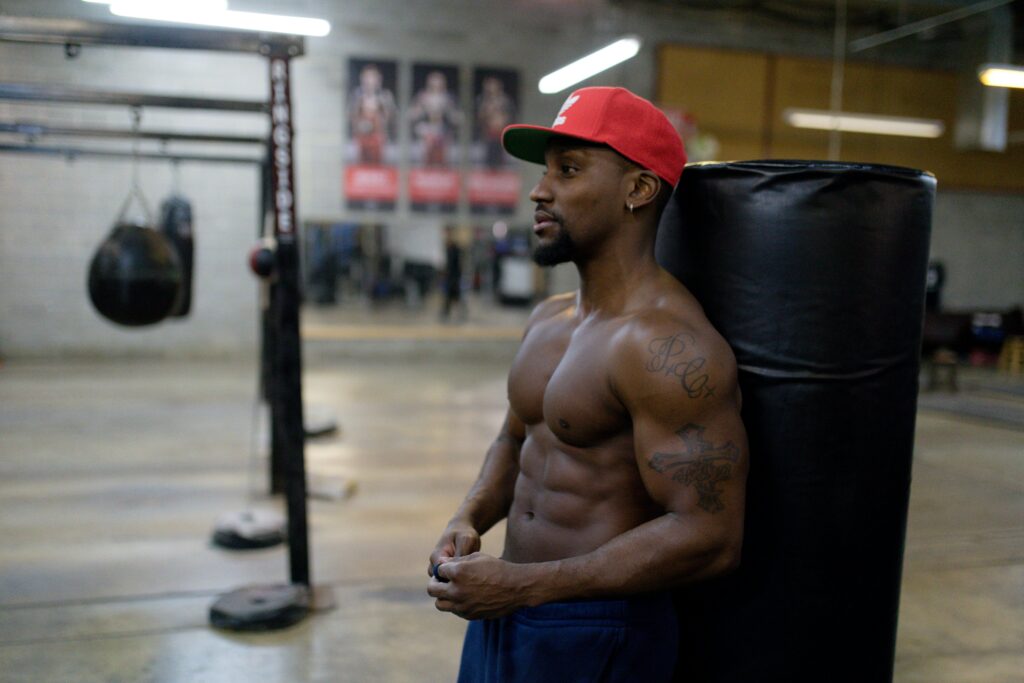 Fit, shirtless man with tattoos leaning on punching bag in a gym setting, exuding strength.